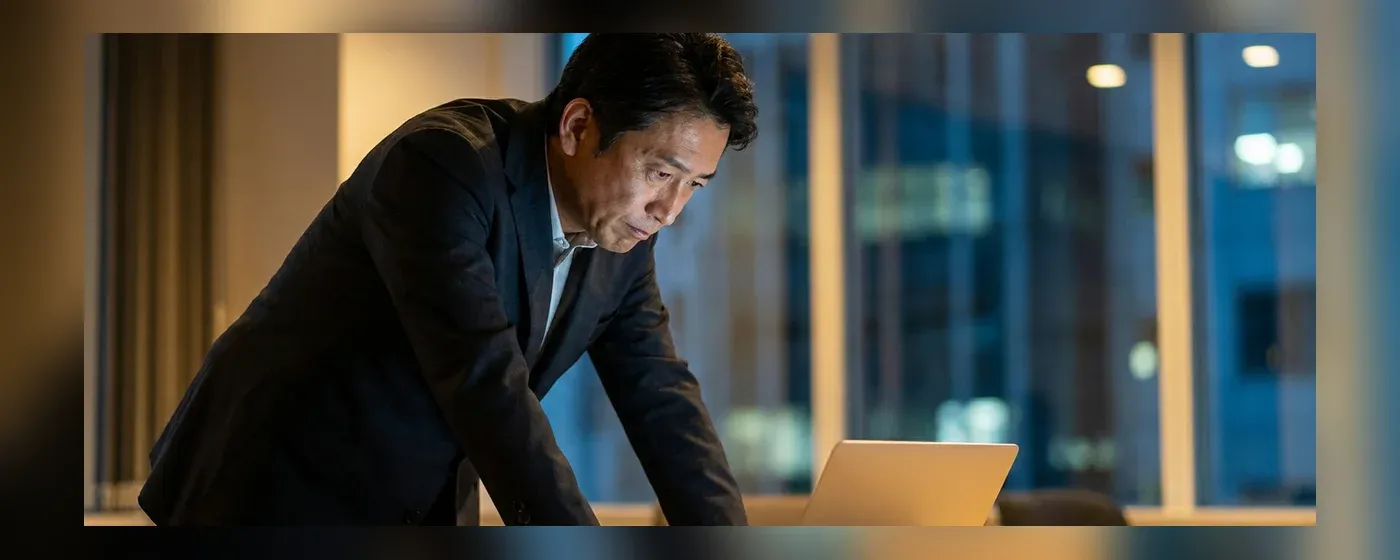 薄暗い部屋でパソコンの画面を見つめながら悩む40代男性ビジネスパーソン